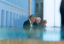 Drei Menschen sitzen auf einem Podium und stellen einen Bericht vor.